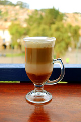 Vertical photo of hot cappuccino coffee in a transparent cup served at the terrace with blurred old town square of Cusco, Peru in background 
