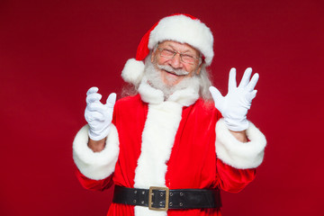 Christmas. Good Santa Claus in white gloves shows faces, grimaces, shows his tongue. Not standard behavior. Isolated on red background.