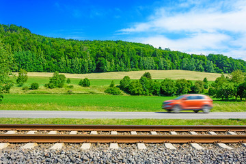 A fast moving car along the road goes alongside the railway tracks. Tracks running next to the road. A sunny view of a car passing through the countryside