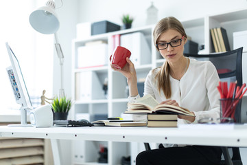 A young girl sits at a table in the office and holds a red cup in her hand and reads a book.