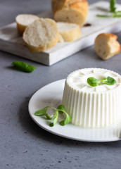Italian ricotta or fresh farm cheese on white plate with corn salad, tomatoes and baguette on grey concrete background. Copy space for text.
