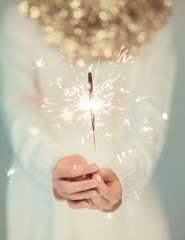 Beautiful woman hands holding sparkler lights, cozy, bright winter sweater with golden christmas garland, can be used as background 