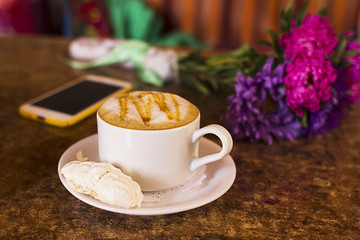 Cup of hot cocoa or Cappuccino or latte coffee with marshmallow and flowers and phone on a table
