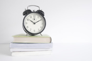 Alarm clock on a pile of books on a wooden shelf. White wall background and empty copy space for Editor's text.