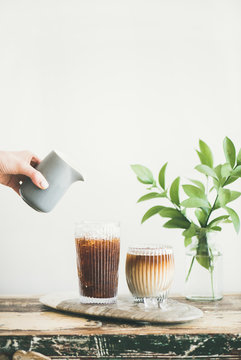 Iced coffee in tall glasses with milk pouring over from pitcher by hand, white wall and green plant branches at background, copy space. Summer refreshing beverage ice coffee concept