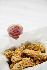 Chicken strips in paper box with sweet chili sauce on a white wooden table, low angle view. Close-up. Copy space.