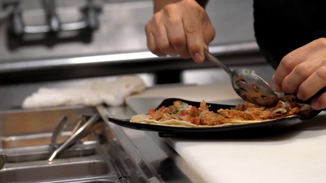 Close up shot of a Hispanic hands carefully topping an order of carnitas tacos with a large spoonful of pico de gallo in the kitchen of a Mexican restaurant