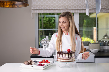 Beautiful female confectioner in chefs uniform standing in a modern kitchen, decorating a naked chocolate and strawberries cake with berries and macaroons