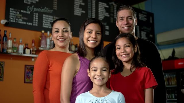 A Beautiful Hispanic Family Gives Each Other A Group Hug While Looking Into The Camera In Their Family Mexican Food Restaurant.