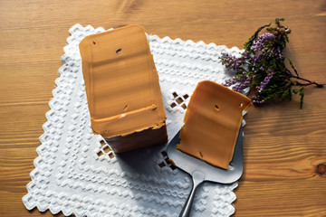 Traditional Norwegian brown cheese on a white napkin with embroidery hardanger. Brunost. Sheep cheese. A piece of soft brown caramel cheese and a slicer on a wooden surface.