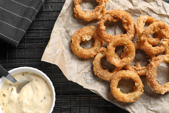 Cooling rack with homemade crunchy fried onion rings and sauce on wooden background, top view