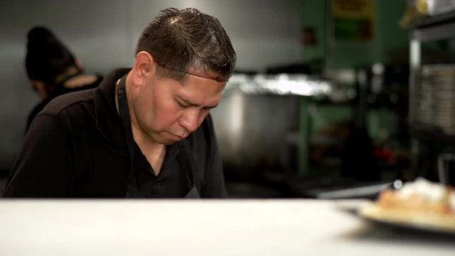 A cook of hispanic descent prepares food in a Mexican food restaurant. He places tacos on the counter and it gets taken away by a server.