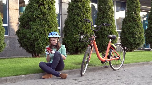 Man and city rolling bicycle, environmentally friendly transport. Beautiful young caucasian woman worker sitting resting on the grass uses a red mobile phone near an orange bicycle with a coryne