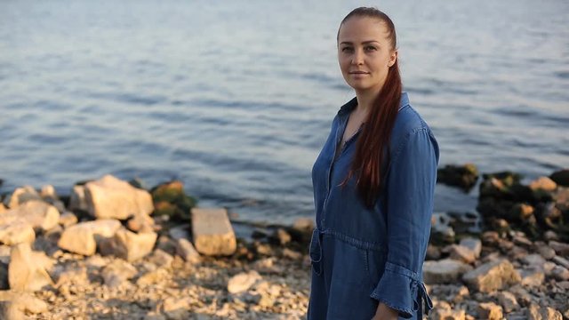 young beautiful Caucasian middle-aged woman with red hair in a blue denim dress stands near the sea on the waterfront in the evening at sunset against the blue sky.