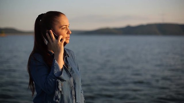 young beautiful Caucasian woman with red hair in the evening at sunset walking along the promenade and talking on the phone.