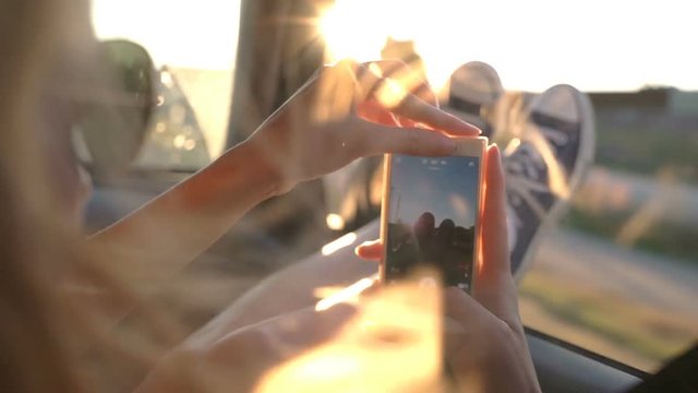 A young redhead woman enjoys traveling in a car by sticking out her legs in an open window and taking pictures using the phone. Slow motion.