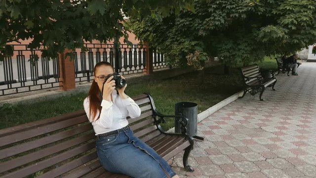 Caucasian middle-aged woman with red hair sitting in the Park on a bench and taking pictures on the camera.