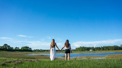 Two women friend blonde & brunette with long hair holding hands in a field next to a lake in florida