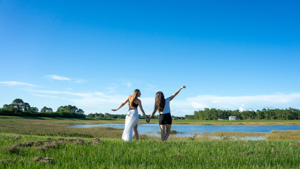 Two women friend blonde & brunette with long hair holding hands in a field and having fun next to a lake in florida
