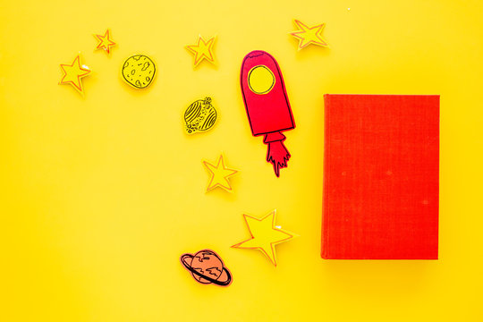 Literature for children. Fantactic, fiction story. Book with blank cover near cutout of rocket, stars, moon on yellow background top view mockup copy space