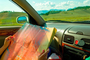 The girl rides in the car in the passenger seat and looks at the paper map looking for the desired route and indicates the road while traveling through the mountains of the Altai