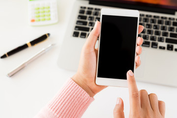 Woman hand holding white mobile phone on a table with a laptop in office.