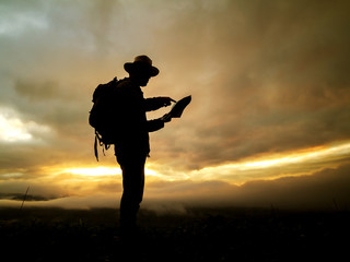 silhouette of travel man with backpack checks map to find directions in wilderness area on mountain at morning sunrise