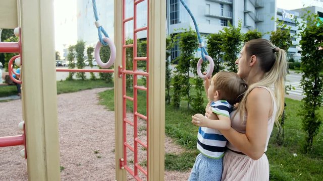 4k video of young mother lifting her toddler son and helping him hanging on rings at park playground