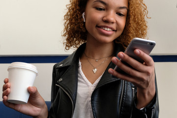 African American girl smiling, looking into her phone. A white glass with coffee in hand. A beautiful young modern black woman, in a leather jacket with airpods in her ear, listens to music.