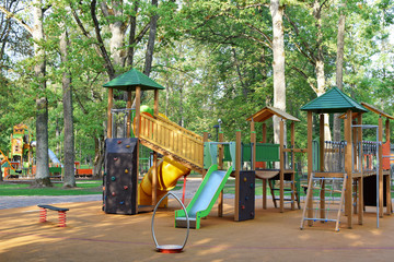 Fototapeta na wymiar Empty colorful playground with swings and slides for children with soft orange protective rubber coating in city park with old trees and pathway in summer day. Children outdoor activities concept.