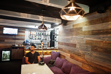 Stylish beard arabian man in glasses and black t-shirt smoking hookah indoor bar. Arab model having rest.
