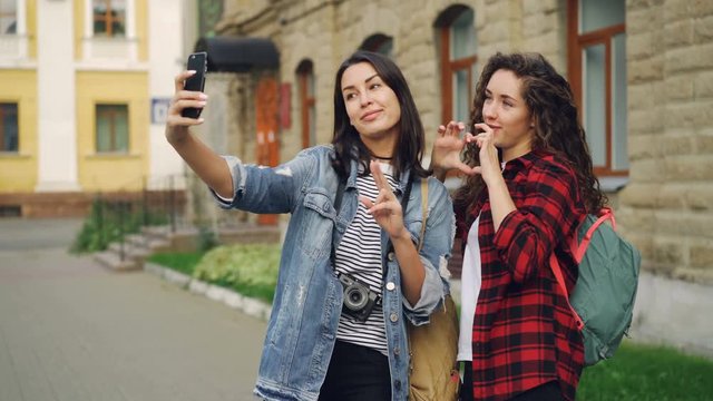 Cheerful girls foreign travelers are taking selfie using smartphone standing outdoors and posing with hand gestures showing v-sign and heart with fingers and laughing.