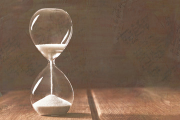 A side view of an hourglass with falling sand, on a dark background with copy space, toned, with faded old letters, a metaphor of time