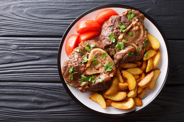 shank Ossobuco with a garnish of fried potato slices and fresh tomatoes close-up on a plate. Horizontal top view