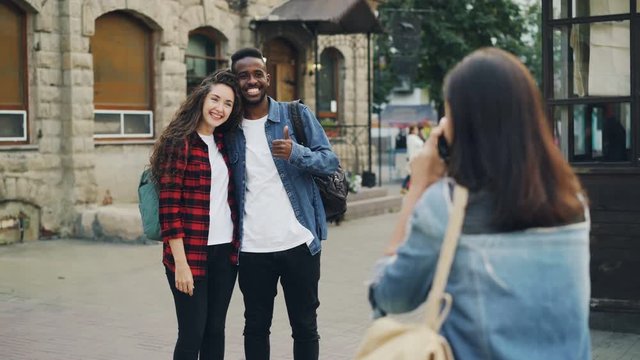 Young woman is taking pictures of beautiful couple African American guy and Caucasian girl standing in the street and posing for camera. Tourism, technology and friends concept.