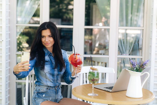 Attractive brunette with long hair, has a European appearance, stylishly dressed in jeans, makes selfie on a smartphone with a glass of grapefruit juice.