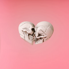 Two skulls in heart shape kissing on pastel pink background. Minimal love concept.