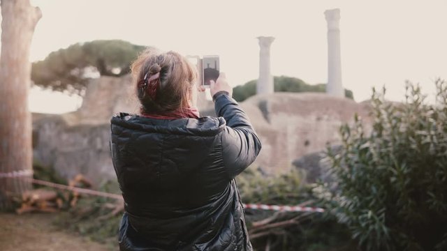 Back view of happy senior smiling Caucasian woman taking a smartphone photo of ancient ruins in Ostia, Italy on vacation