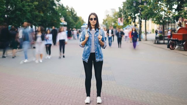 Time-lapse of attractive young woman wearing sunglasses standing alone in big city with backpack and looking at camera when crowds of people are passing by.
