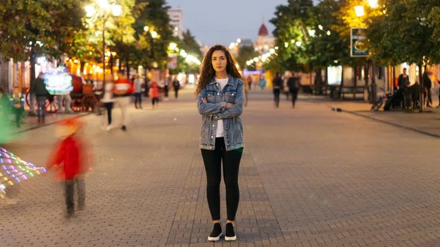 Time-lapse of attractive woman with long curly hair standing in the street with crossed arms and looking at camera on autumn evening when people are moving around.