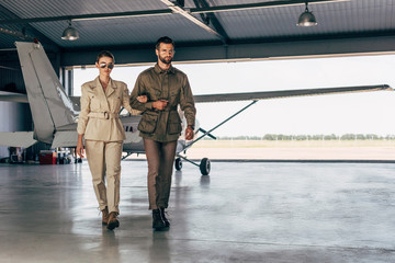 confident fashionable young couple in jackets walking near airplane in hangar - obrazy, fototapety, plakaty