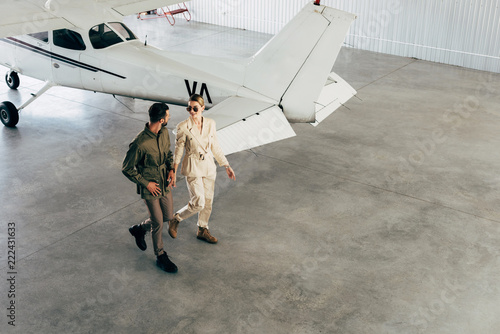 high angle view of young couple in stylish jackets walking in hangar with airplane