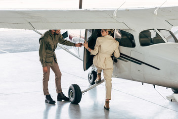 stylish young man opening door for girlfriend while she boarding in plane