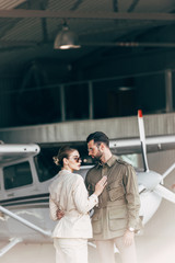  fashionable young couple in stylish jackets standing in hangar with airplane