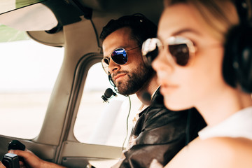 selective focus of handsome male pilot in headset and sunglasses sitting with girlfriend in cabin of airplane - obrazy, fototapety, plakaty