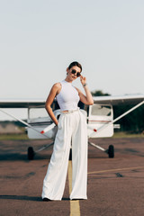 stylish young woman in sunglasses posing near airplane