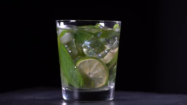 Lemon fruit Caipirinha alcoholic beverage of Brazil in glass on black background, close up. Rotates cold drink with green mint leaf, lime and ice, macro