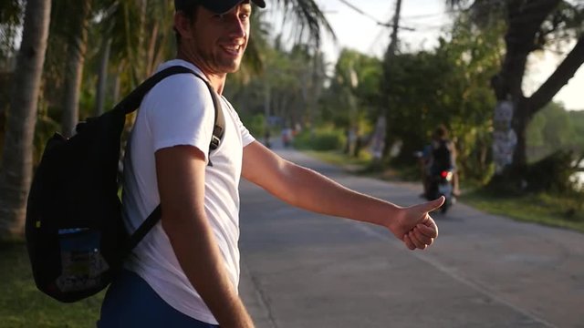 A young cheerful man with a backpack behind him, catches a car hitchhiking, raised his thumb. slow motion, HD, 1920x1080