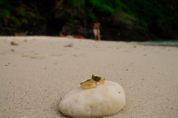Two golden rings on a piece of coral with a young couple kissing at the beach in the background