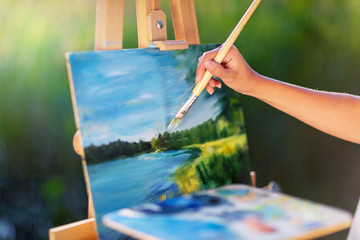 Obraz premium Young pretty woman artist draws paints a picture of a lake on open plain air outdoors